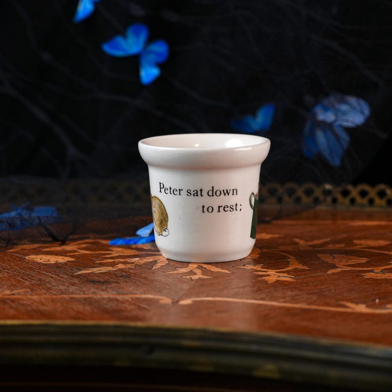 Op de afbeelding: Een witte keramische beker met een bruine konijnillustratie en de zwarte tekst Peter sat down to rest; aan de zijkant. De beker heeft een uitlopende rand en een gladde afwerking, met een klassiek verhaalkarakter op de buitenkant.