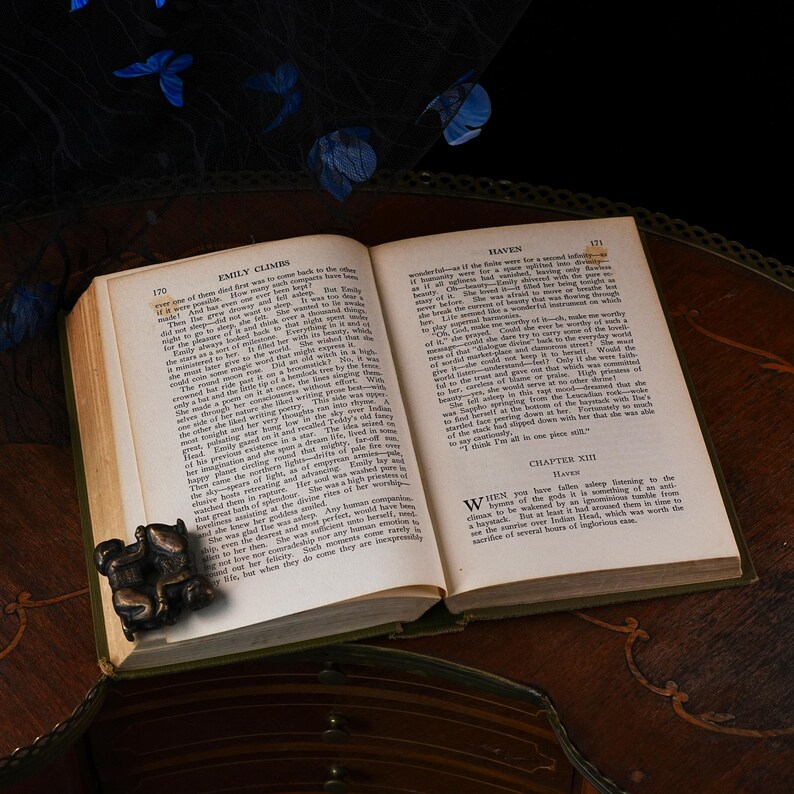Puede incluir: Un libro antiguo de tapa dura abierto en las p&aacute;ginas 170 y 171, con texto impreso en negro. Una peque&ntilde;a escultura de metal en tono bronce oscuro que representa una figura sentada est&aacute; colocada en la esquina inferior izquierda de las p&aacute;ginas.