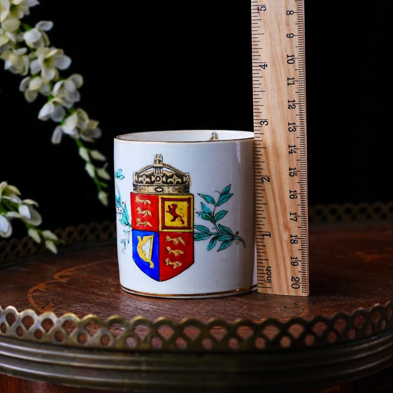 May include: A white ceramic mug with a gold rim, featuring a royal crest design with a crown and shield. The mug is approximately 3 inches tall, as measured by a wooden ruler. Decorative floral elements are also present.