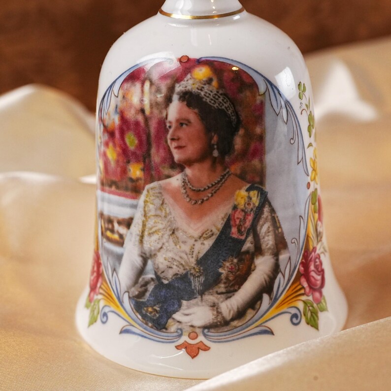 K&ouml;nnte beinhalten: Wei&szlig;e Keramikglocke mit einem Portr&auml;t einer Frau mit Tiara und Sch&auml;rpe. Die Glocke ist mit floralen und verzierten Bord&uuml;ren in Rot, Blau und Gelb verziert. Sie hat einen goldenen Rand.