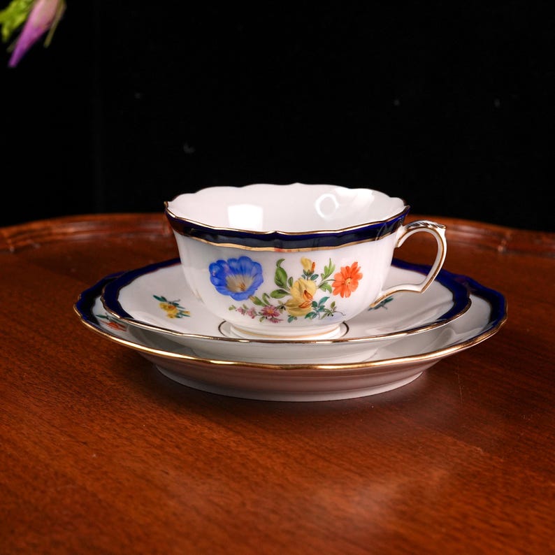 Puede incluir: Juego de taza y platillo de porcelana blanca con borde azul marino y detalles dorados. La taza y el platillo est&aacute;n decorados con motivos florales coloridos. El juego est&aacute; sobre una superficie de madera.