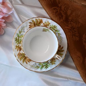 Op de afbeelding: Een witte theekop en schotel met een gouden rand en een decoratief bladerpatroon in groen en bruin. De theekop staat op de schotel, tegen een witte satijnen achtergrond.