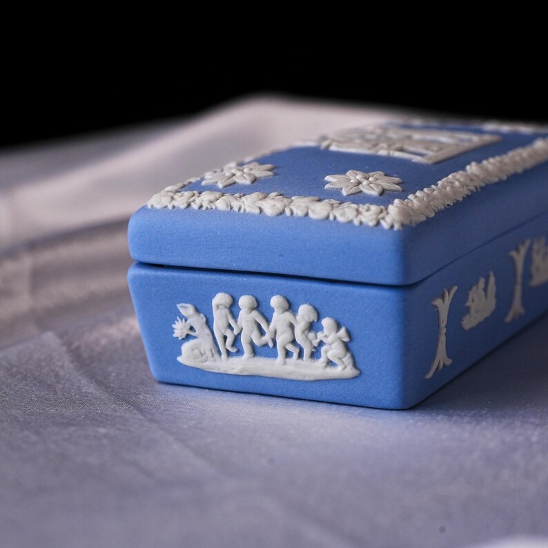 May include: A blue rectangular trinket box with white relief detailing. The box features floral and classical figures in relief. The box is a light blue color and has a rectangular shape. The box is likely made of ceramic.