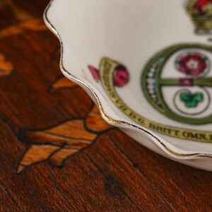 Op de afbeelding: Een witte porseleinen schaal met een geschulpte rand afgewerkt met goud. De binnenzijde bevat een decoratief embleem met een groene letter E, een roze roos, een groen klaverblad en een geel lint met de tekst BRITT OMN REX F D.
