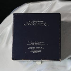 Może przedstawiać: Ciemnoniebieskie pudełko z białym tekstem. Tekst zawiera "&copy; 1995 Royal Doulton", "Royal Doulton&reg; is a trademark" i tekst francuski. Dodatkowy tekst wskazuje producenta i importera, w tym adres w Scarborough, Ontario.