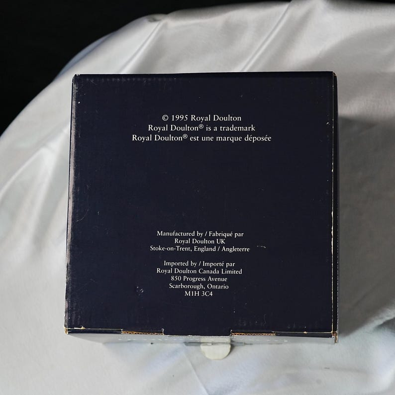 Może przedstawiać: Ciemnoniebieskie pudełko z białym tekstem. Tekst zawiera "&copy; 1995 Royal Doulton", "Royal Doulton&reg; is a trademark" i tekst francuski. Dodatkowy tekst wskazuje producenta i importera, w tym adres w Scarborough, Ontario.