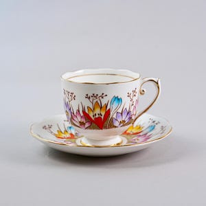 May include: A white teacup and saucer set with a floral design. The cup and saucer have a gold trim and feature colorful flowers in shades of red, pink, yellow, blue, and purple. The teacup has a curved handle.