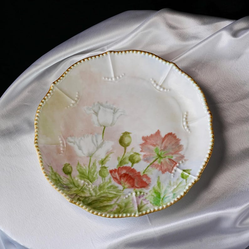 May include: A white porcelain plate with a scalloped edge and a gold beaded rim. The plate features hand-painted red and white poppy flowers with green leaves and buds. The background has a soft pink hue.