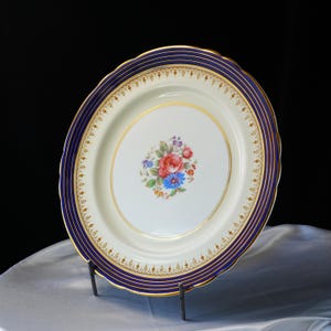 Pode incluir: Travessa oval para servir com um centro creme e uma borda azul marinho e dourada. O centro apresenta um buqu&ecirc; floral de flores vermelhas, azuis e roxas. A travessa &eacute; suportada por um suporte de metal preto.
