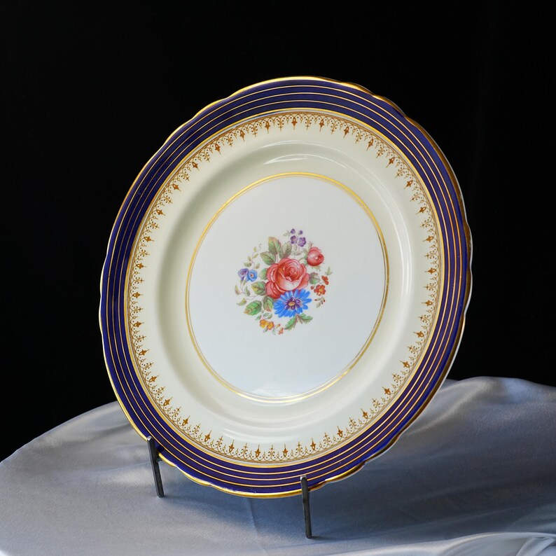 Pode incluir: Travessa oval para servir com um centro creme e uma borda azul marinho e dourada. O centro apresenta um buqu&ecirc; floral de flores vermelhas, azuis e roxas. A travessa &eacute; suportada por um suporte de metal preto.