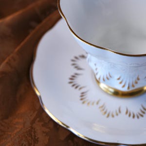 May include: White teacup and saucer set with gold trim. The teacup has a scalloped rim and a decorative pattern. The saucer has a matching scalloped edge and gold accents. The set is on a brown surface.