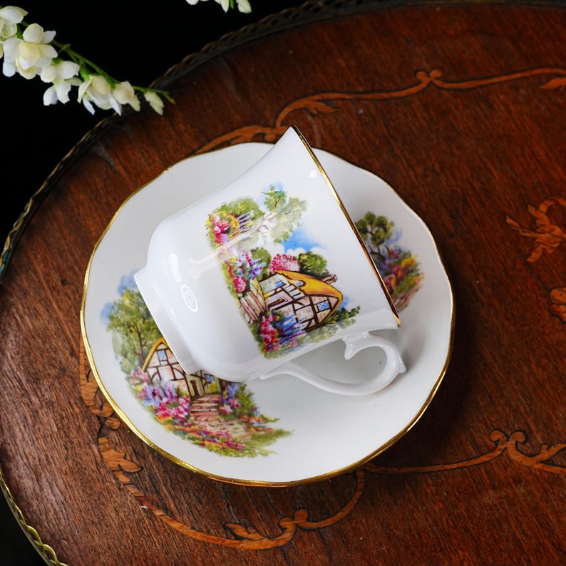 May include: White teacup and saucer set with a gold rim. The cup and saucer feature a colorful cottage scene with flowers and trees. The set is placed on a dark wooden surface.