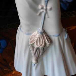 K&ouml;nnte beinhalten: Eine gl&auml;nzende wei&szlig;e Porzellanfigur mit einem langen, flie&szlig;enden Rock und einem hellblauen Banddetail. Ein kleiner, skulptierter Sonnenschirm in zarten Rosa- und Blaut&ouml;nen ist an der Vorderseite des Rocks befestigt und mit einer zarten Porzellanschleife verziert.