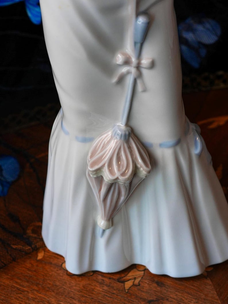 K&ouml;nnte beinhalten: Eine gl&auml;nzende wei&szlig;e Porzellanfigur mit einem langen, flie&szlig;enden Rock und einem hellblauen Banddetail. Ein kleiner, skulptierter Sonnenschirm in zarten Rosa- und Blaut&ouml;nen ist an der Vorderseite des Rocks befestigt und mit einer zarten Porzellanschleife verziert.