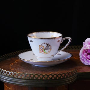 May include: A white teacup and saucer set with gold edging. The teacup has a colourful emblem with the letter "R" and a bird. The saucer and cup have a delicate design. The set is on a wooden surface.
