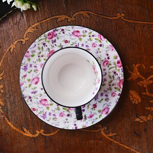 May include: A white teacup and saucer set featuring a floral design of pink roses and green foliage. The cup has a black handle and rim, and the saucer has a matching black rim. The set is on a dark wooden surface.