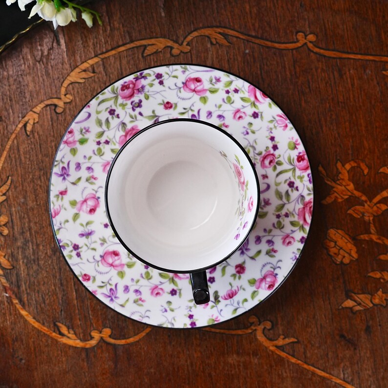 May include: A white teacup and saucer set featuring a floral design of pink roses and green foliage. The cup has a black handle and rim, and the saucer has a matching black rim. The set is on a dark wooden surface.