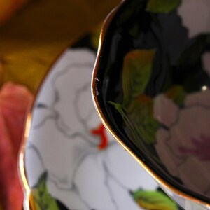 May include: Close-up of a porcelain teacup and saucer set. The teacup is black with a gold rim and floral design. The saucer is white with a matching floral pattern. The flowers are white with green leaves and a touch of red.