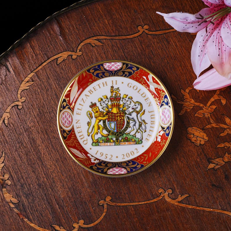 May include: A round porcelain commemorative plate featuring the Royal Coat of Arms of the United Kingdom in the center. The text reads Queen Elizabeth II Golden Jubilee 1952-2002. The border is decorated with red, blue, and gold floral patterns.