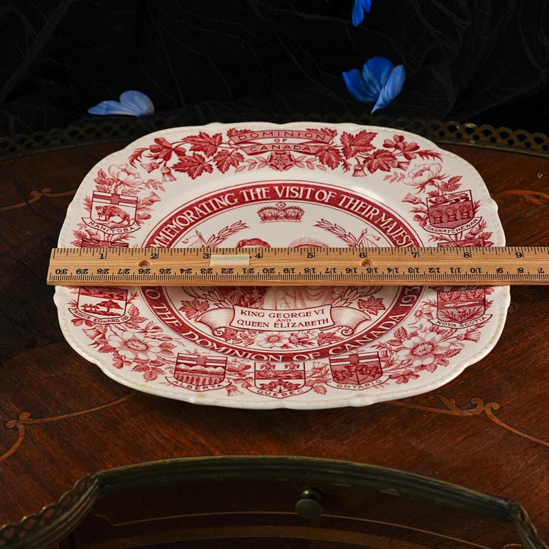 May include: A white ceramic commemorative plate with red transferware patterns, featuring portraits of King George VI and Queen Elizabeth. A wooden ruler placed across the center shows the plate measures approximately 26.7 cm in width.