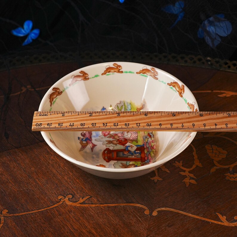 May include: A cream-colored ceramic bowl featuring a repeating pattern of brown rabbits along the inner rim and a central illustration of rabbits near a red telephone box. A wooden ruler shows the bowl has a diameter of approximately 6 inches.