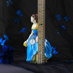 May include: A Royal Doulton porcelain figurine of a person in a blue dress with a yellow shawl, holding yellow flowers. A wooden ruler placed next to the figurine shows a height of approximately 8.5 inches from the base to the top of the head.