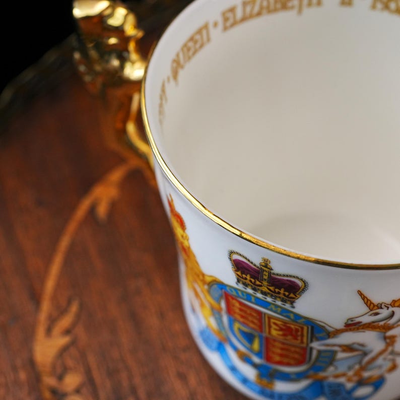 Puede incluir: Una taza de porcelana blanca con borde dorado y asa dorada. El exterior muestra un colorido escudo de armas real con una corona, un le&oacute;n y un unicornio. El borde interior tiene la inscripci&oacute;n QUEEN ELIZABETH II.