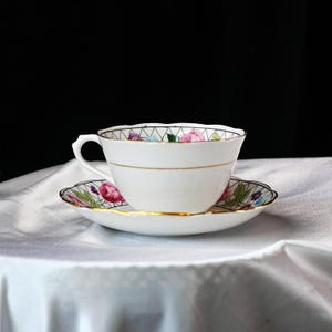 Op de afbeelding: Een witte kop en schotel set met een bloemenontwerp. De kop en schotel hebben een gouden rand. Het bloemenpatroon bevat roze, paarse en groene bloemen. De kop heeft een gebogen handvat.