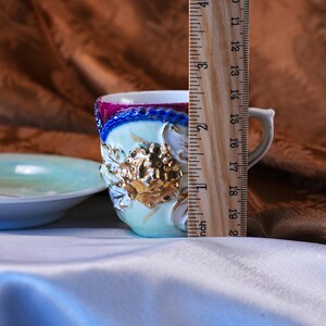 May include: A vintage teacup and saucer set. The teacup is approximately 2 inches tall, with a light blue and gold floral design. The rim is trimmed with a dark pink and blue border. A wooden ruler is placed next to the teacup.