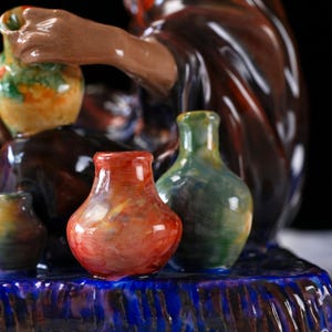 May include: A selection of small, hand-painted ceramic vases in shades of red, green, and yellow. The vases are displayed on a blue and brown decorative base, with a figure in the background. The style is artistic and detailed, with a focus on colour.