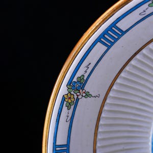 May include: A detailed shot of a decorative plate with a gold rim and a white interior. The plate has a blue and gold border with a floral design in blue, yellow, and white. The plate has a ridged texture.