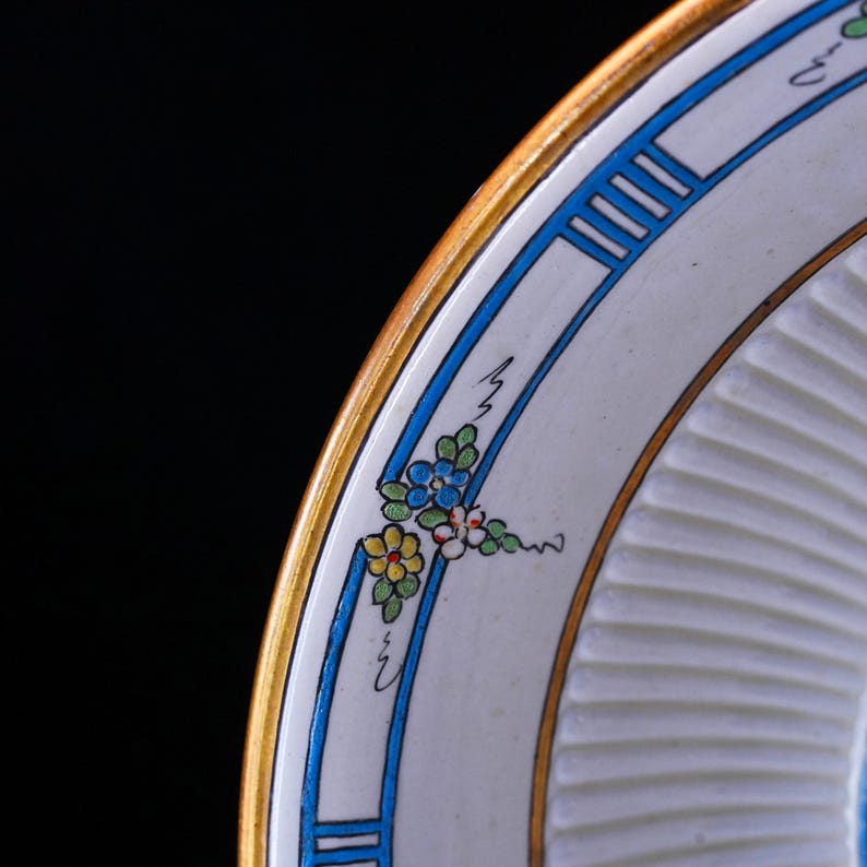 May include: A detailed shot of a decorative plate with a gold rim and a white interior. The plate has a blue and gold border with a floral design in blue, yellow, and white. The plate has a ridged texture.