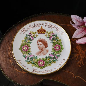 Op de afbeelding: Een wit keramieken herdenkingsbord met een centraal portret van koningin Elizabeth II met een kroon. Het portret is omringd door een bloemenkrans van rozen, distels en klaverbladeren. De tekst luidt H.M. Queen Elizabeth II, June 2nd, 1953, Coronation.