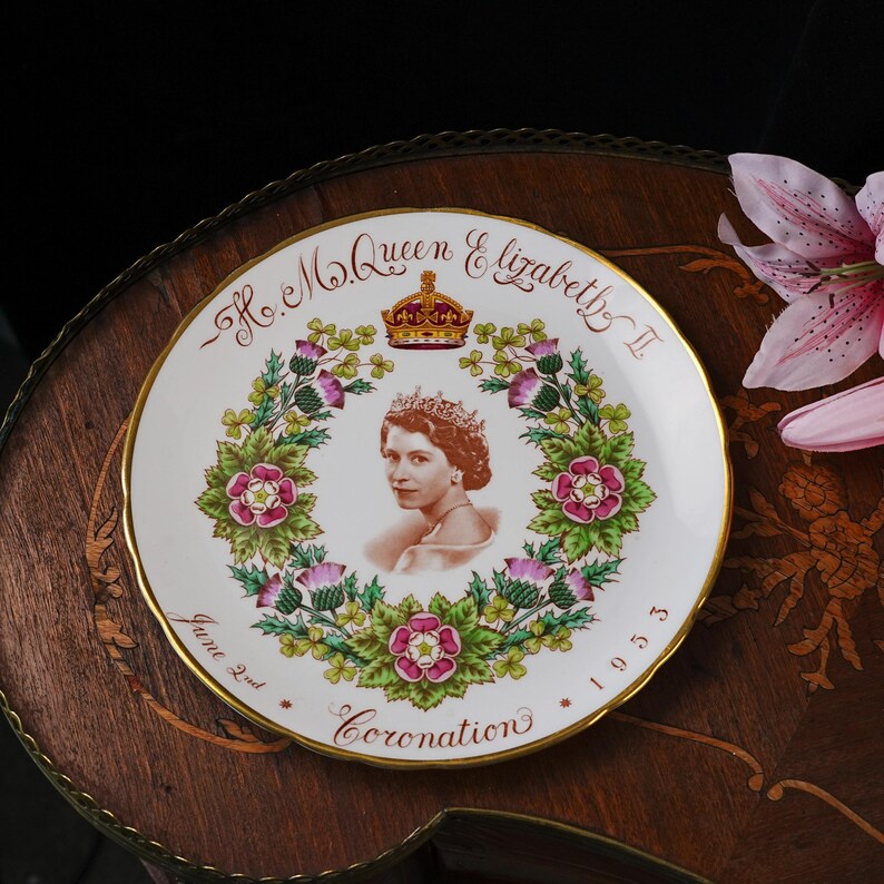 Op de afbeelding: Een wit keramieken herdenkingsbord met een centraal portret van koningin Elizabeth II met een kroon. Het portret is omringd door een bloemenkrans van rozen, distels en klaverbladeren. De tekst luidt H.M. Queen Elizabeth II, June 2nd, 1953, Coronation.