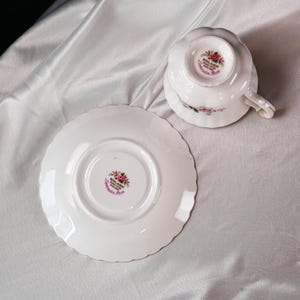以下が含まれることがあります： 花柄の白いティーカップとソーサーのセット。ティーカップは逆さまになっており、底のフローラルパターンが見えます。ソーサーとティーカップはスカラップエッジで、「Royal Albert Bone China England Lavender Rose」の文字がソーサーとティーカップの底に印刷されています。