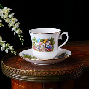 May include: White teacup and saucer set with a gold rim. The cup and saucer feature a colorful cottage scene with a yellow roof and vibrant flowers. The set is placed on a wooden surface.
