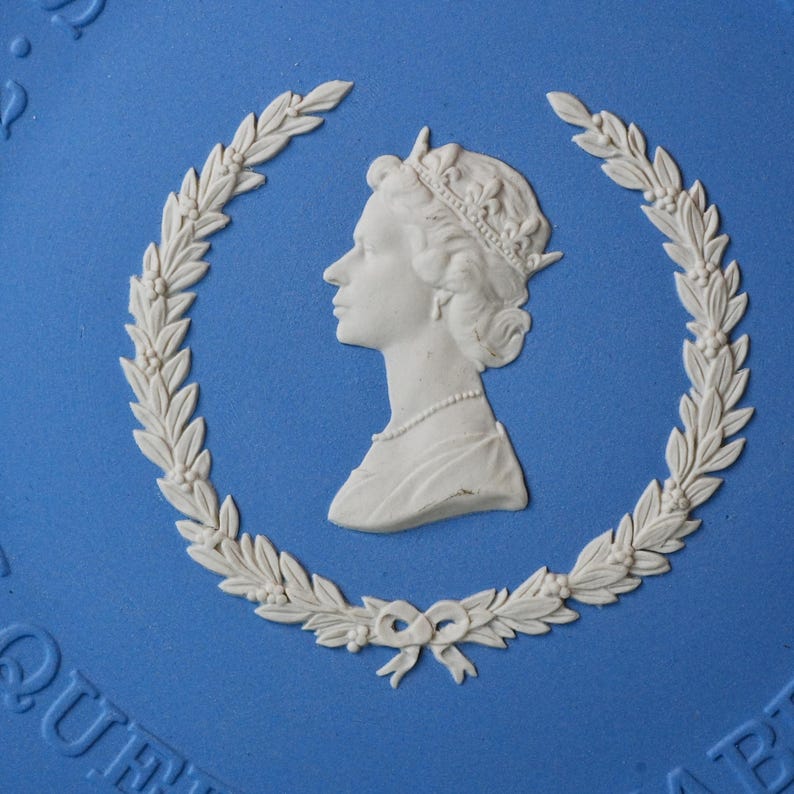 May include: A blue ceramic plate with a white relief of a profile portrait inside a laurel wreath. The portrait shows a woman wearing a crown and a necklace. The plate has a glossy finish.