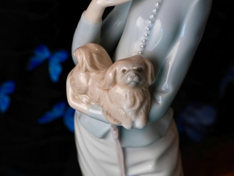 K&ouml;nnte beinhalten: Eine gl&auml;nzende Porzellanfigur, die eine Person in einer hellblauen Jacke mit einer vertikalen Reihe kleiner wei&szlig;er Kn&ouml;pfe zeigt, die einen kleinen, hellbraunen, langhaarigen Hund mit Schlappohren und glatter Oberfl&auml;che h&auml;lt.