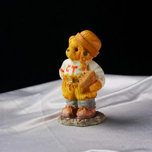 Op de afbeelding: Een beeldje van een teddybeer in een voetbaluniform, compleet met helm en bal. De outfit van de beer is wit met oranje details en de letters "CT". Hij staat op een kleine, getextureerde basis. Het kleurenschema is warm.