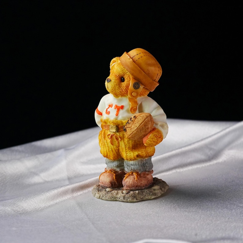Op de afbeelding: Een beeldje van een teddybeer in een voetbaluniform, compleet met helm en bal. De outfit van de beer is wit met oranje details en de letters "CT". Hij staat op een kleine, getextureerde basis. Het kleurenschema is warm.