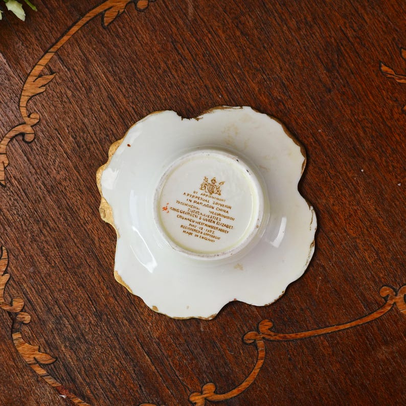 Op de afbeelding: Een witte en gouden porseleinen schotel met een geschulpte rand. Het midden heeft een cirkelvormige inkeping en tekst. De schotel staat op een houten oppervlak. De tekst bevat de woorden "By Appointment" en "Made in England."