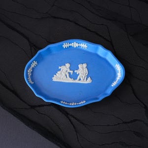 May include: An oval-shaped blue ceramic tray featuring a raised white relief design of two figures in the centre. The scalloped edges of the tray are decorated with white floral and leaf motifs positioned at the top, bottom, and sides.