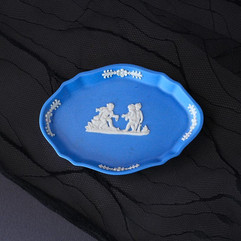 May include: An oval-shaped blue ceramic tray featuring a raised white relief design of two figures in the centre. The scalloped edges of the tray are decorated with white floral and leaf motifs positioned at the top, bottom, and sides.