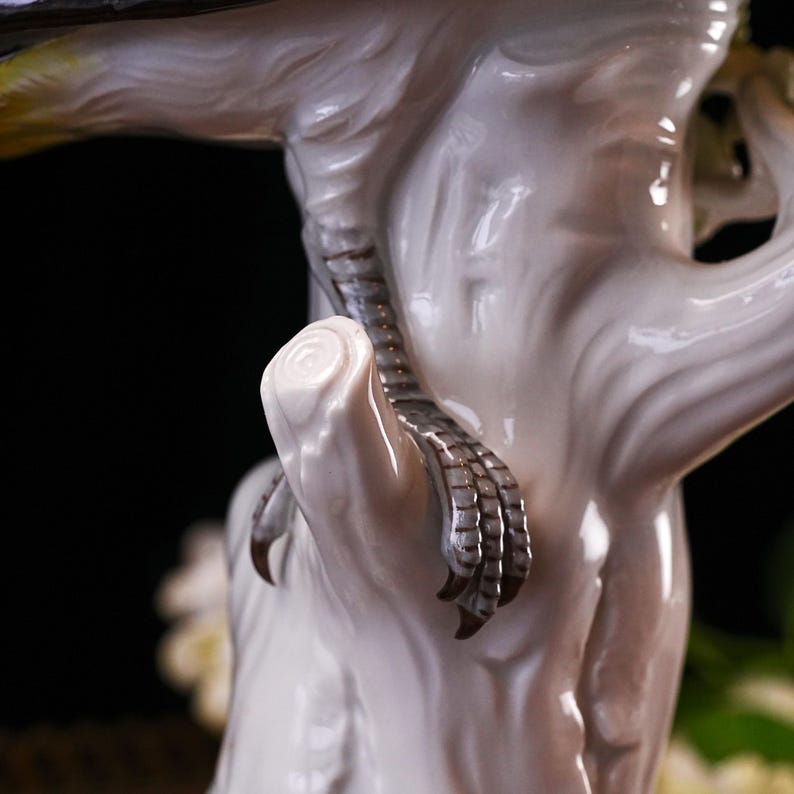 May include: A detailed close-up of a white ceramic bird figurine perched on a tree branch. The bird's clawed foot is visible, with detailed texture and color. The branch is also white, with subtle texture. The background is dark.