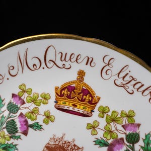 Op de afbeelding: Een wit porseleinen bord met een goudkleurige rand en de tekst M. Queen Elizabeth in bruin schrift. Het ontwerp bevat een centrale afbeelding van een koninklijke kroon in goud, rood en paars, geflankeerd door groene en paarse distelbloemen.