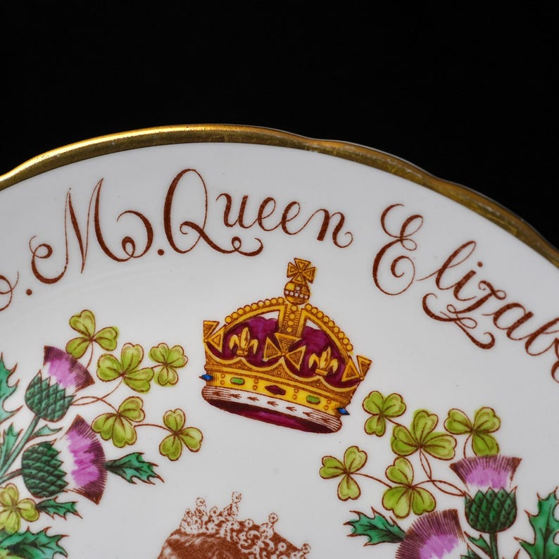 Op de afbeelding: Een wit porseleinen bord met een goudkleurige rand en de tekst M. Queen Elizabeth in bruin schrift. Het ontwerp bevat een centrale afbeelding van een koninklijke kroon in goud, rood en paars, geflankeerd door groene en paarse distelbloemen.