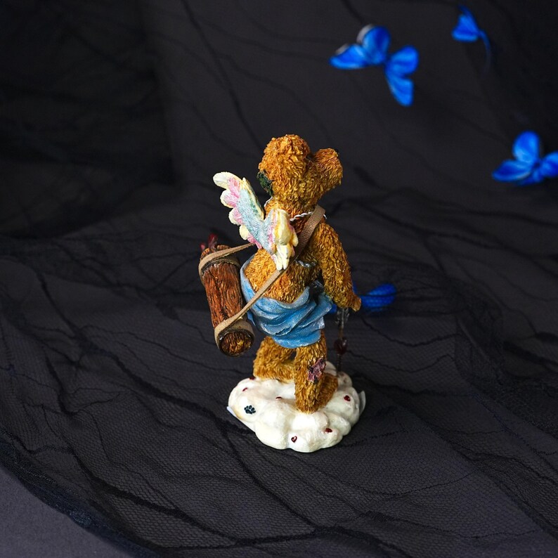 Puede incluir: Una figura de oso de peluche marr&oacute;n sobre una base de nube blanca. El oso tiene pelaje texturizado, alas multicolores y una tela azul alrededor de la cintura. Lleva un objeto cil&iacute;ndrico marr&oacute;n en la espalda con una correa y sostiene un objeto azul.