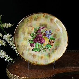 May include: A round decorative plate with a gold rim. The plate features a hand-painted design of various fruits, including purple raspberries, blackberries, and a yellow pear, set against a textured green and yellow background. The plate is displayed on a metal stand.
