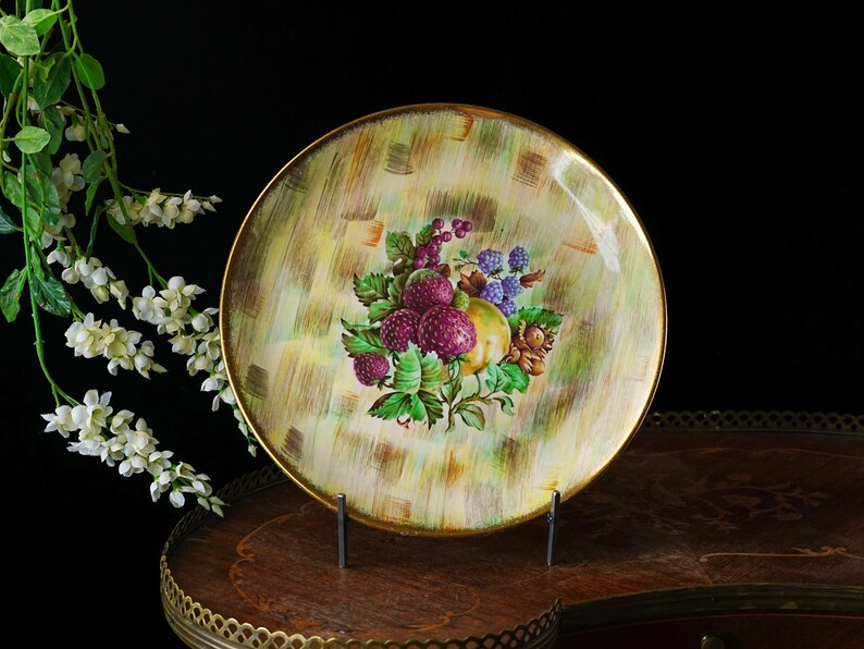 May include: A round decorative plate with a gold rim. The plate features a hand-painted design of various fruits, including purple raspberries, blackberries, and a yellow pear, set against a textured green and yellow background. The plate is displayed on a metal stand.