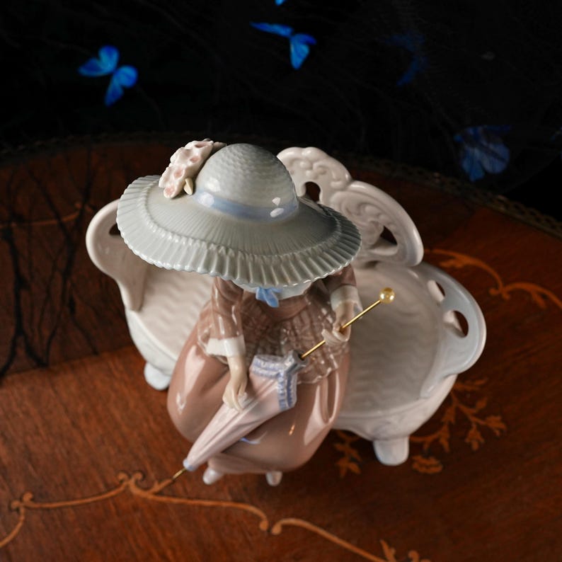 K&ouml;nnte beinhalten: Eine Porzellanfigur einer sitzenden Person mit einem hellblauen Hut mit breiter Krempe und Blumenakzent, einem braunen karierten Oberteil und einem braunen Rock. Die Figur h&auml;lt einen geschlossenen hellblau-wei&szlig;en Sonnenschirm mit Goldspitze.